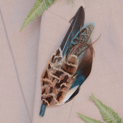 A decorative statement feather boutonniere pin with brown, black, and blue patterned feathers and a teal cord wrapped stem, pinned to a light beige suit jacket with fern leaves as props.