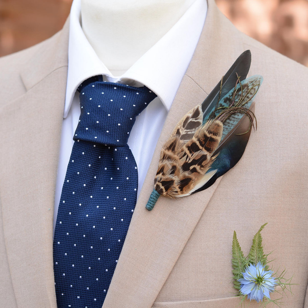 Men's formal wear featuring a patterned feather boutonniere pin with brown, black, iridescent blue and teal feathers, styled on a beige suit jacket, white dress shirt, and navy blue polka dot tie. A small blue flower and fern leaves are tucked in the pocket.