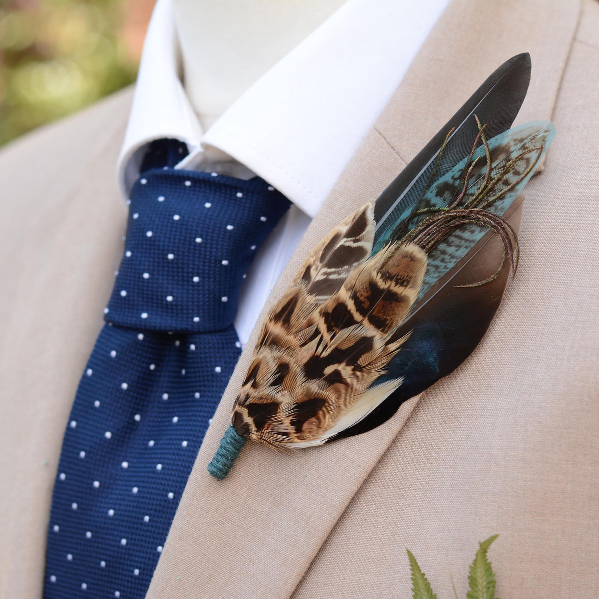 A close-up of men's formal wear accessories  featuring a patterned feather boutonniere pin with brown, black, iridescent blue and teal feathers, styled on a beige suit jacket, white dress shirt, and navy blue polka dot tie.