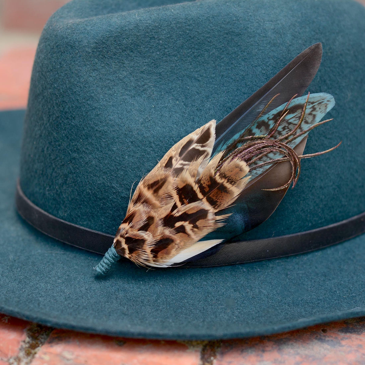 A dark teal felt hat with a black band, decorated with a  feather hat pin with patterned brown and black hen pheasant feathers, a iridescent blue duck feather and teal feathers resting on a brick surface.