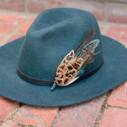 A dark teal felt hat with a black band, decorated with a  feather hat pin with patterned brown and black hen pheasant feathers, a iridescent blue duck feather and teal feathers resting on a brick surface.