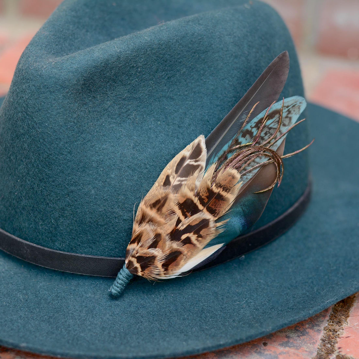 A dark teal felt hat with a black band, decorated with a  feather hat pin with patterned brown and black hen pheasant feathers, a iridescent blue duck feather and teal feathers resting on a brick surface.