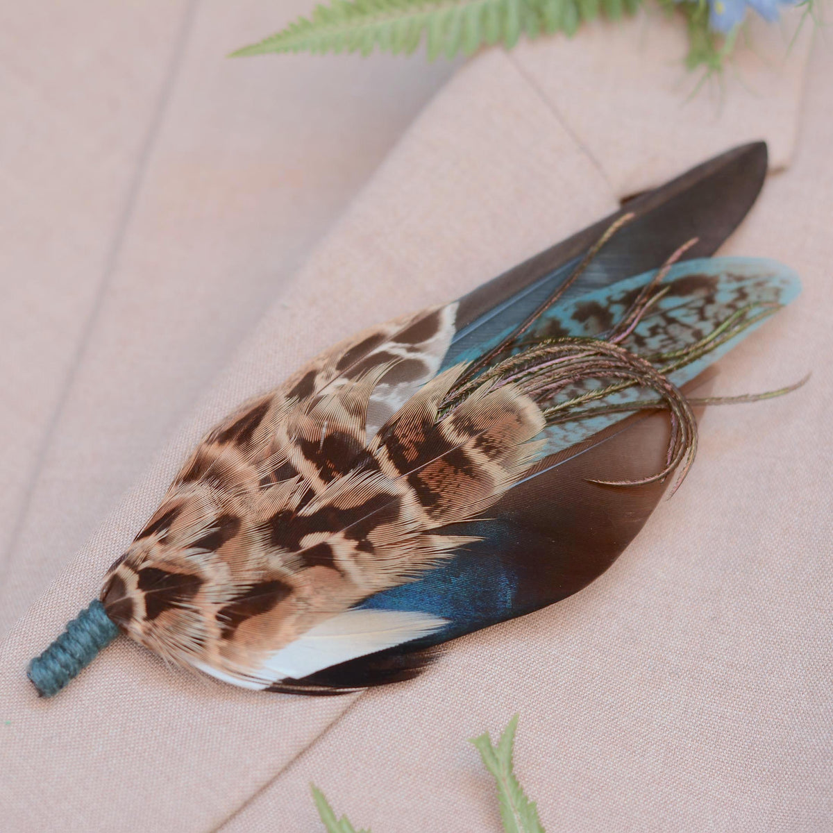 A decorative feather brooch pin with a mottled brown and black pattern, a blue iridescent duck feather, teal green feathers, and a teal twine wrapped stem, photographed on a soft beige fabric surface with small green leaves nearby.