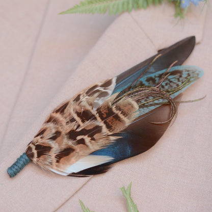A decorative feather brooch pin with a mottled brown and black pattern, a blue iridescent duck feather, teal green feathers, and a teal twine wrapped stem, photographed on a soft beige fabric surface with small green leaves nearby.