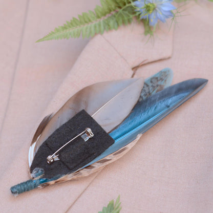 A decorative feather pin with a silver safety pin clasp and felt backing, displayed on a beige fabric surface with a fern and small blue flower.