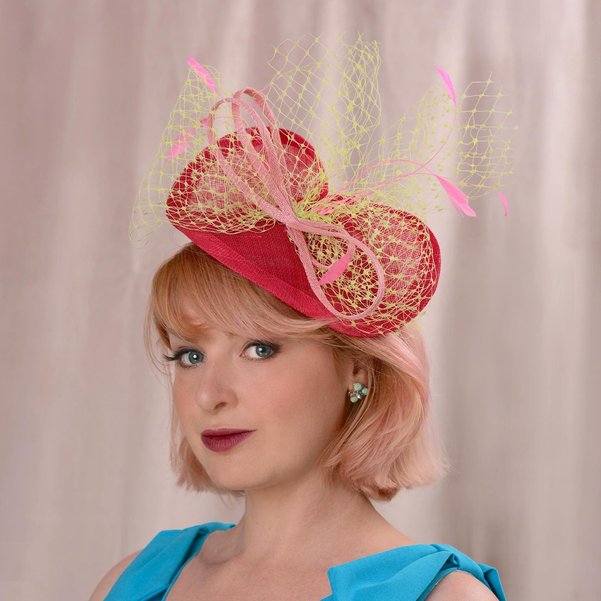 A woman with short pink hair wears a bright pink fascinator hat with a yellow net veiling bow and tonal pink details, posed against a soft beige curtain background.