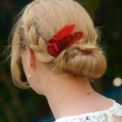 A woman with blonde hair in a bridal plaited bun up do decorated with a red and black feather hair clip with striped and spotted markings