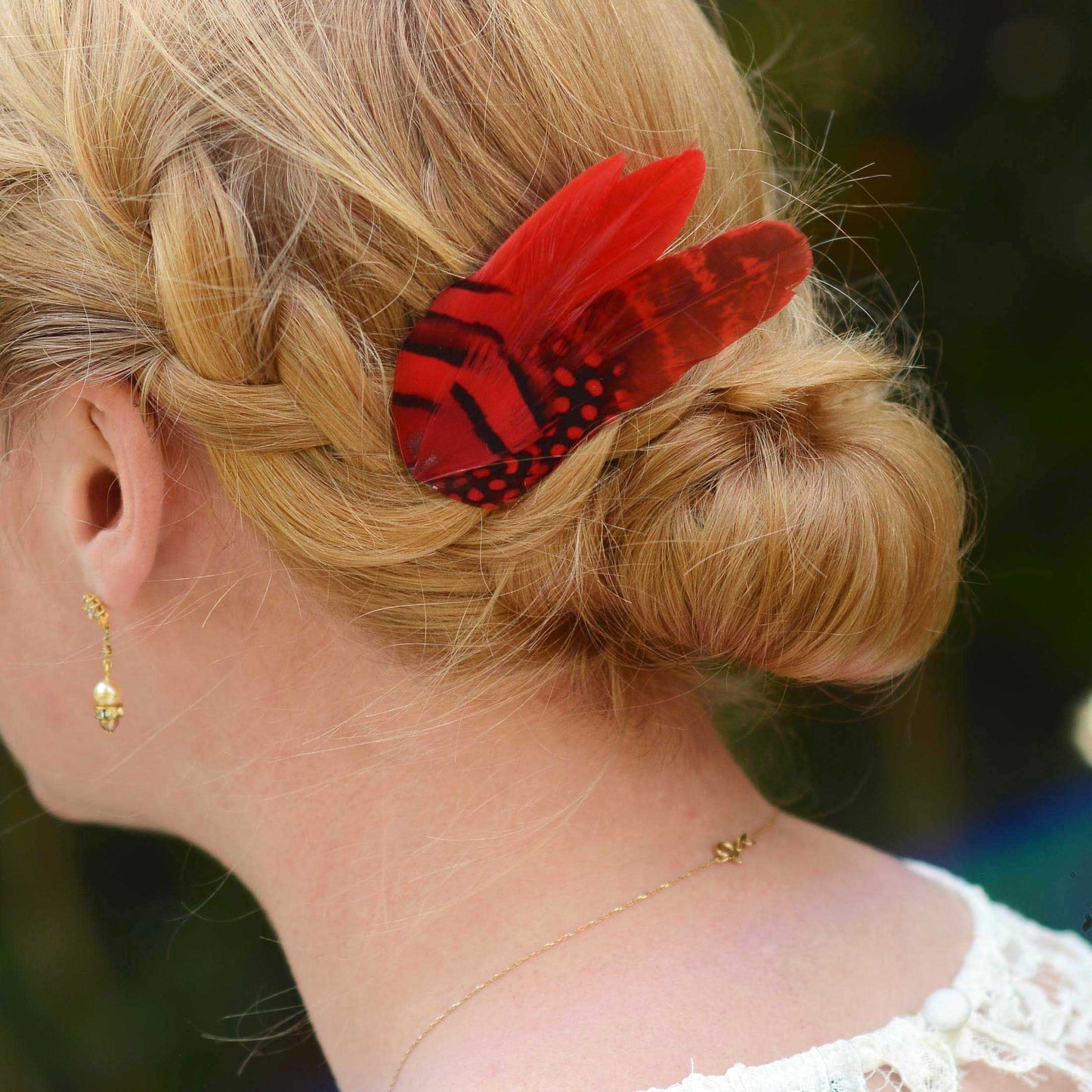 A woman with blonde hair in a formal plaited bun up do decorated with a red and black feather hair clip with striped and spotted markings