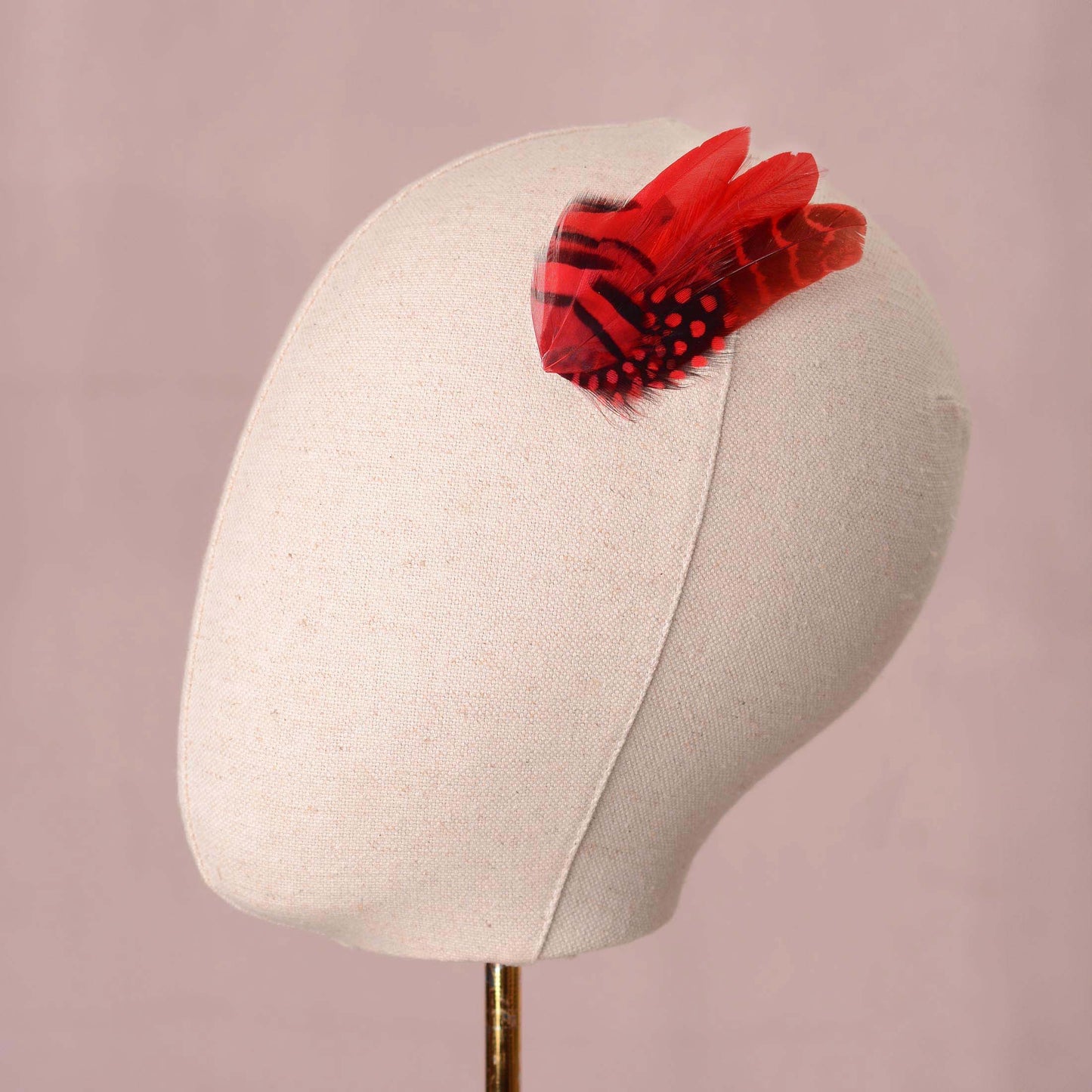 A red and black patterned feather hair accessory displayed on a cream fabric mannequin head against a soft pink background.