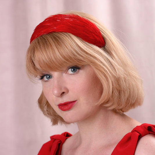A woman with blonde bob-cut hair wearing a textured red headband covered in fine feathers and a red dress, photographed against a soft pink curtain background.