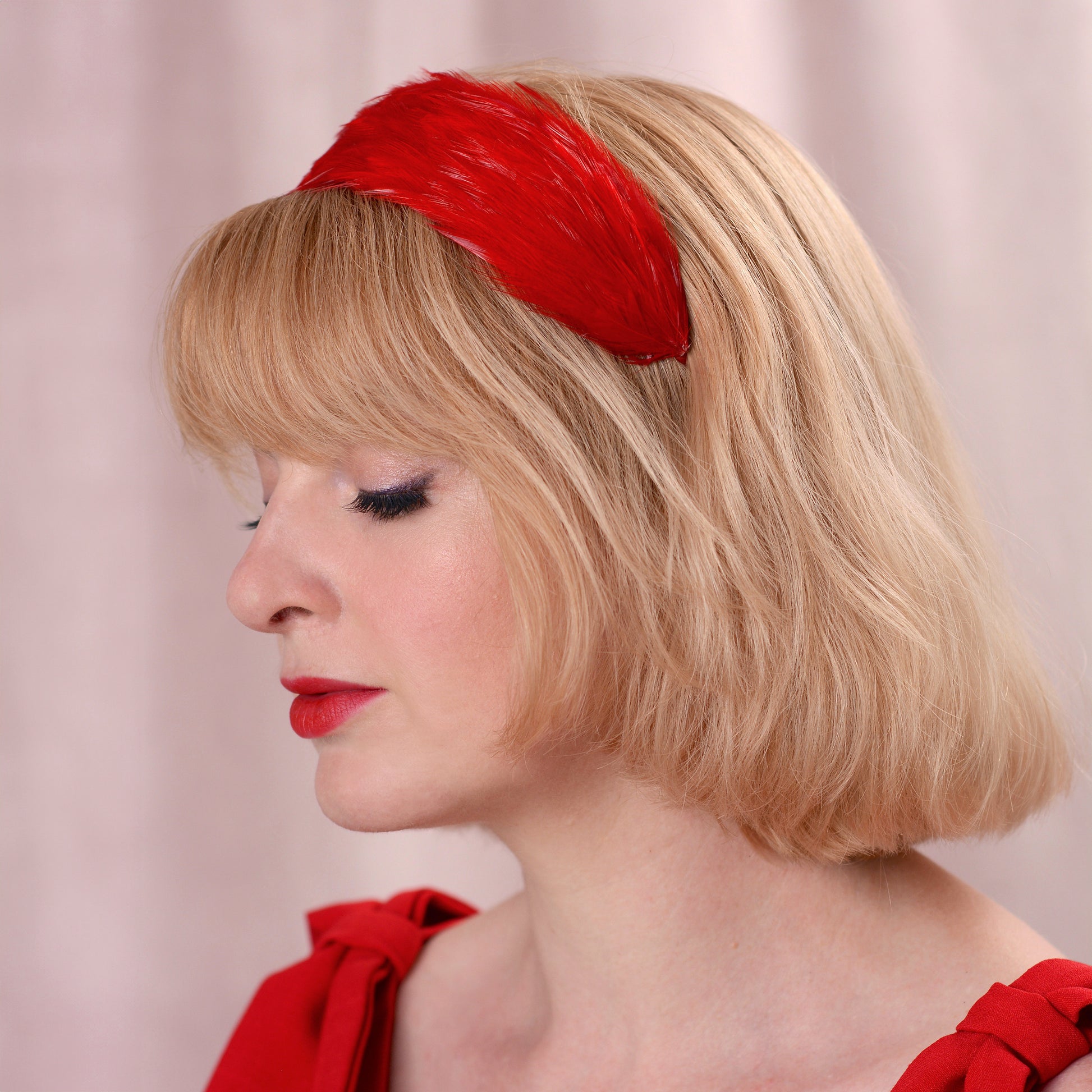 A woman with a short blonde bob wearing a bright red feather headband, shown in profile against a soft pink background.