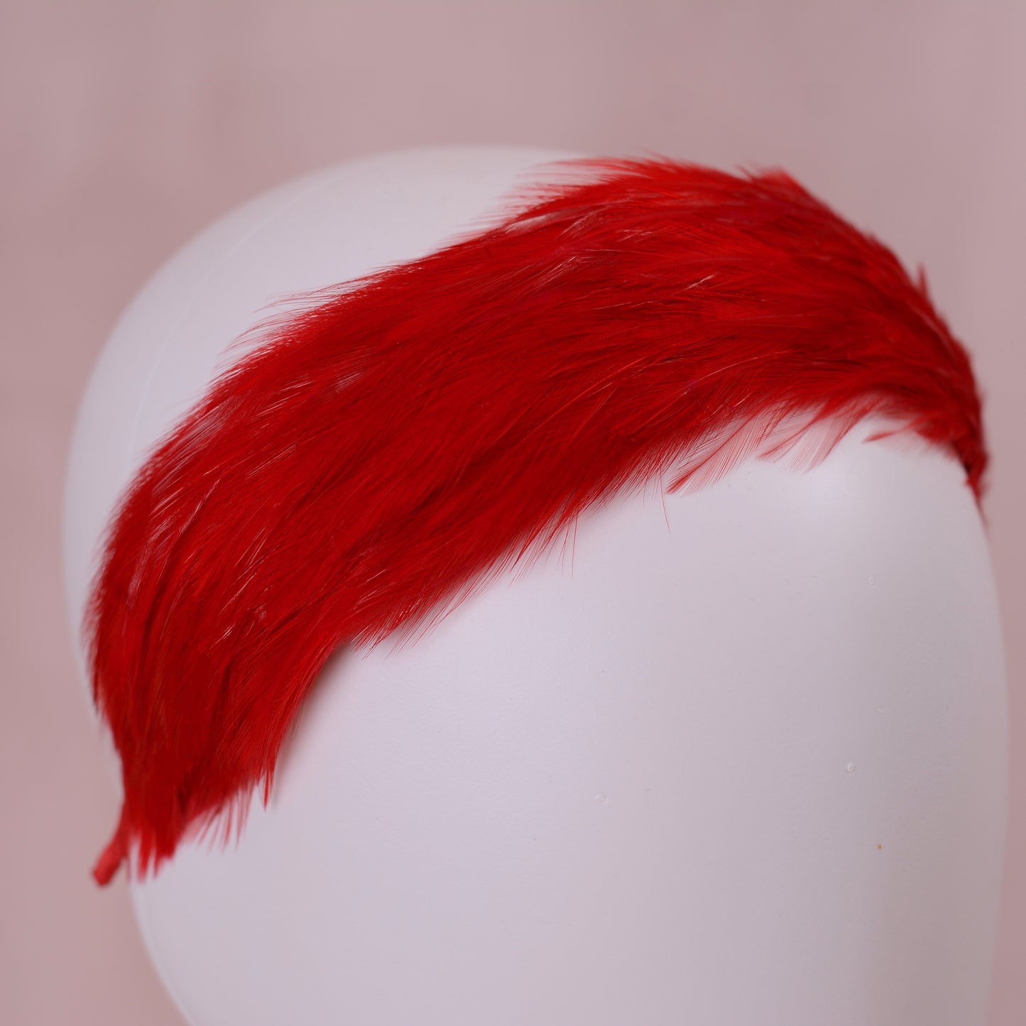 A bright red feather headband made with hundreds of fine rooster feathers displayed on a white mannequin head against a soft pink background.