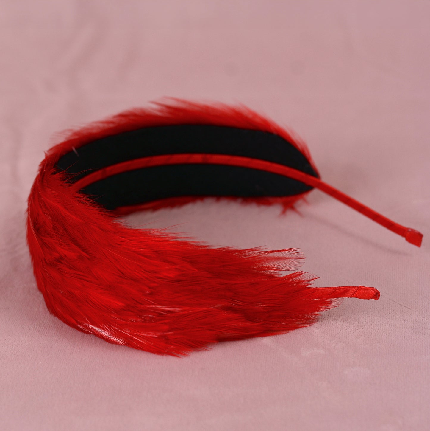 A red feather aliceband with a black inner lining and a red metal headband, displayed on a light pink fabric background.