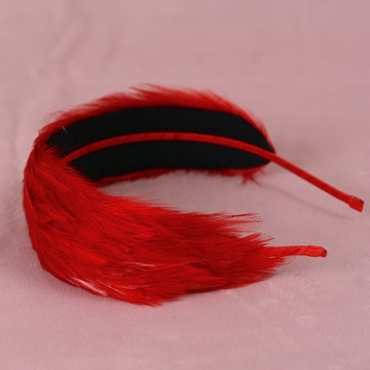 A red feather aliceband with a black inner lining and a red metal headband, displayed on a light pink fabric background.