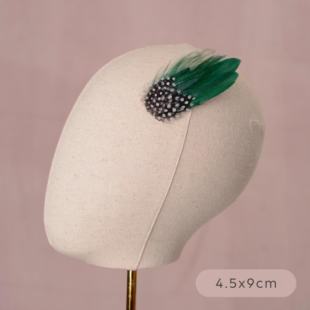 A decorative hair accessory displayed on a beige fabric mannequin head against a soft pink background. The accessory features green feathers paired with polka-dot guinea fowl feathers.
