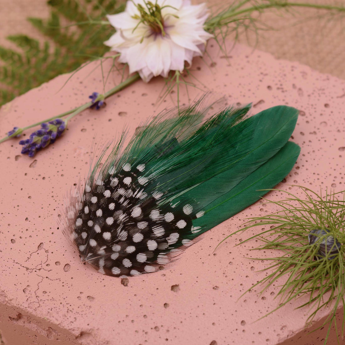 A decorative feather hair accessory featuring black and white spotted feathers and emerald green feathers, displayed on a textured pink stone surface with floral props.
