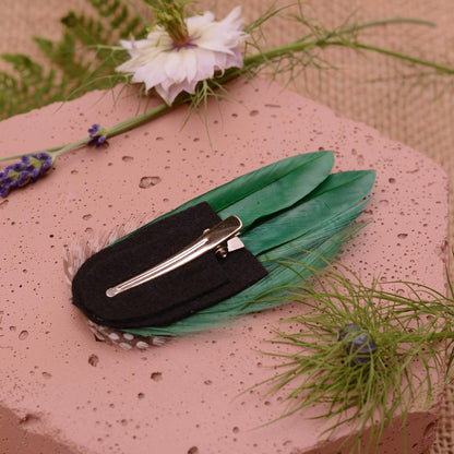 A decorative green feather hair clip featuring a black felt base and silver-toned alligator-style clip, displayed on a pink textured stone with floral props.