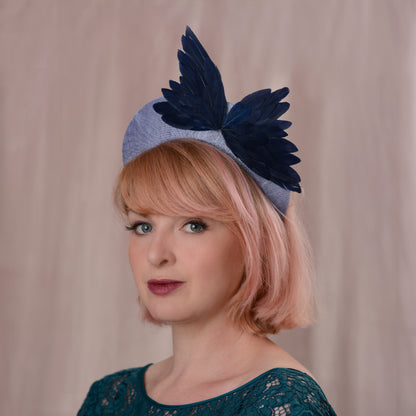 A woman with pink hair wearing a light blue straw fascinator hat in a bandeau headband shape with dark navy feather bird wings, set against a soft beige curtain background.