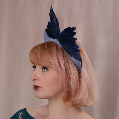 A woman with pink hair wearing a navy blue feather fascinator headpiece on a light blue headband, photographed against a soft beige backdrop.