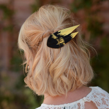 A blonde woman shown from the back wearing a black and gold feather hair clip in her hair, with a soft-focus outdoor background.