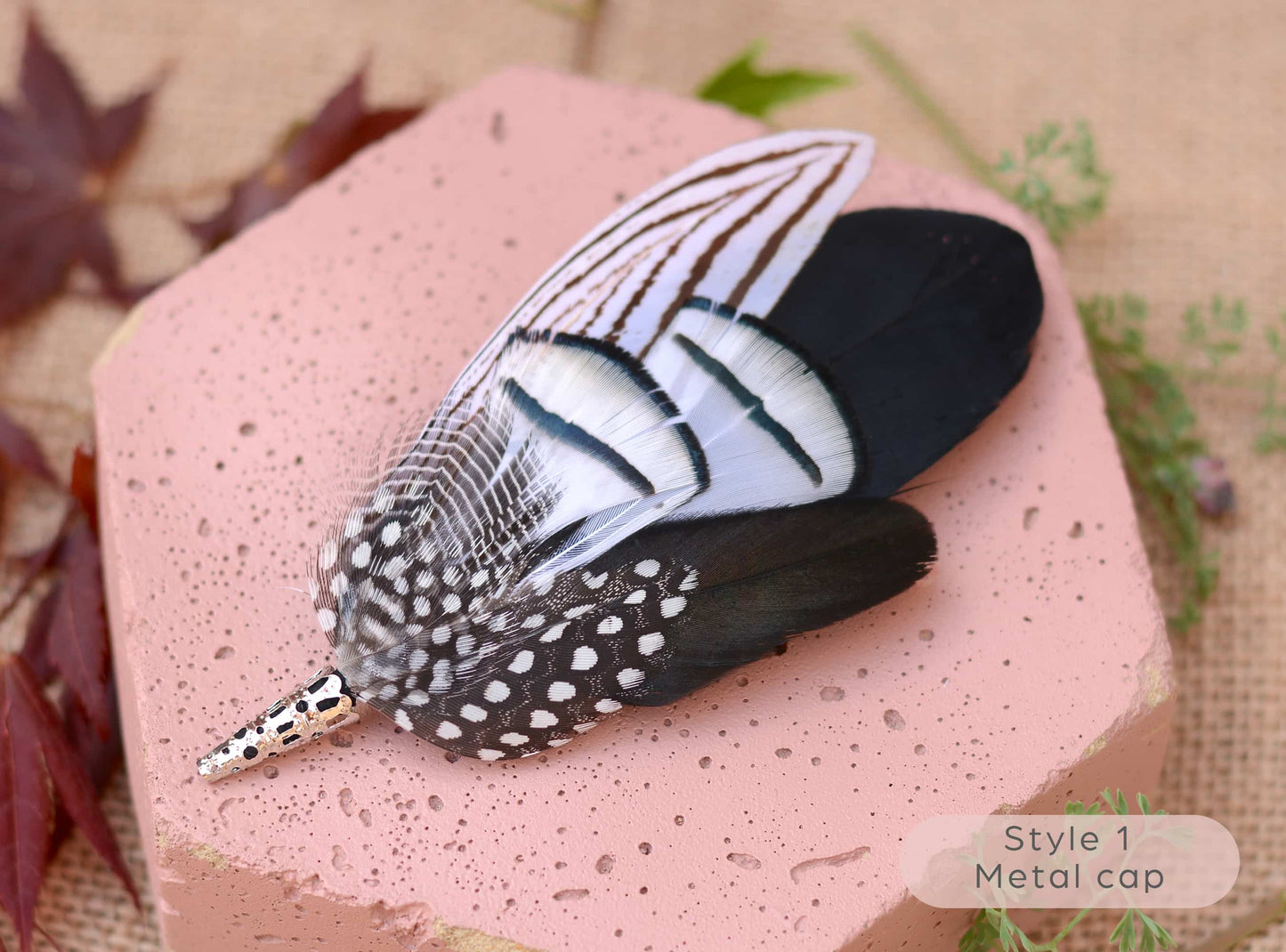 Decorative large feather boutonniere with a metal cap on a pink stone surface with leaves and grass in the background.