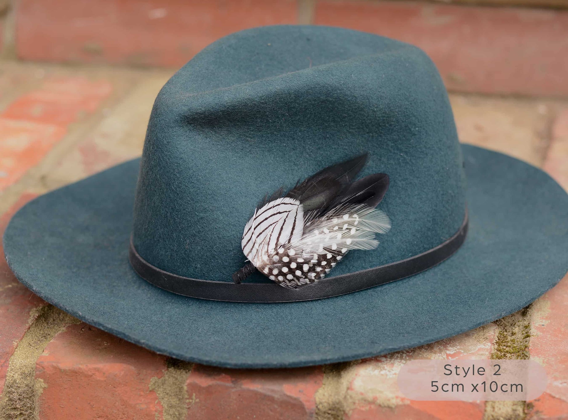 Teal fedora hat with black and white feather hat pin detail on a brick surface