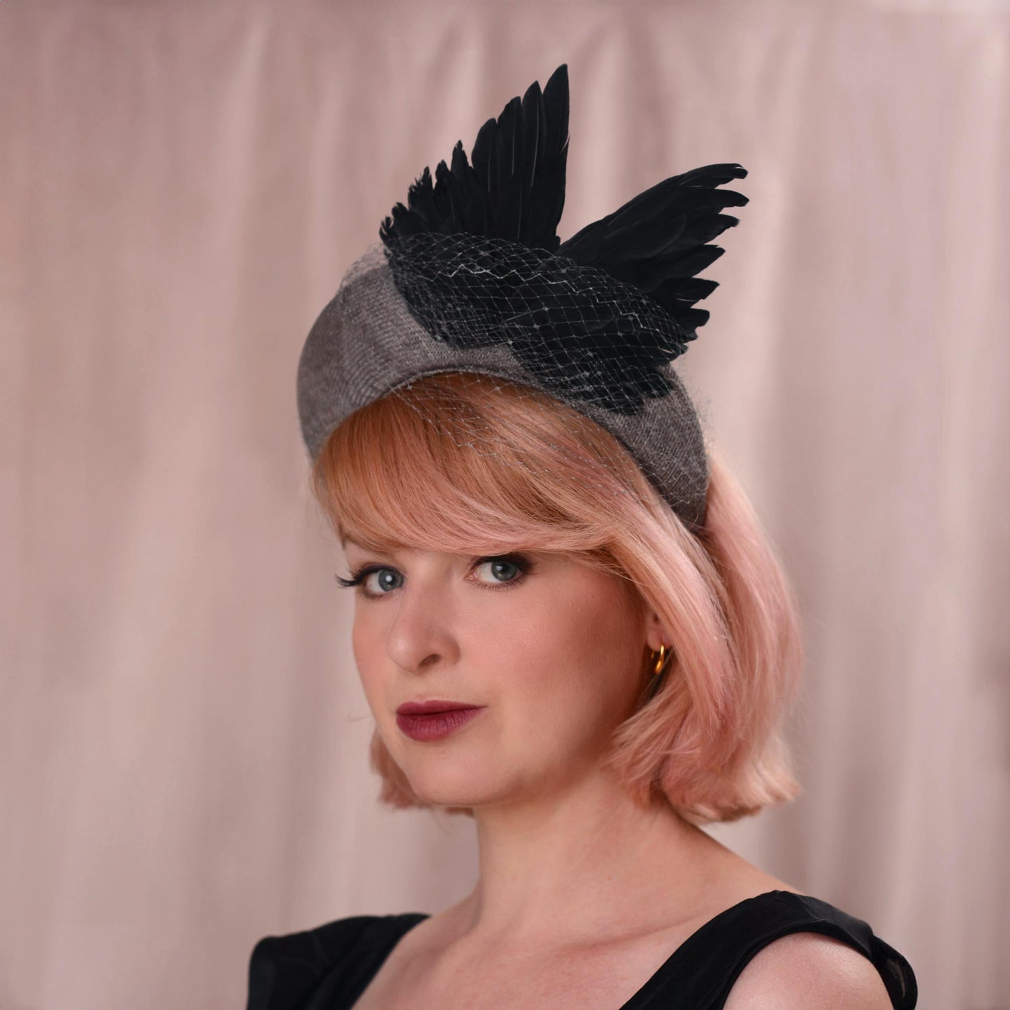 A woman with short pink hair wears a black bird wing fascinator hat with a grey headband base and fine net veiling, posed against a soft beige curtain backdrop.