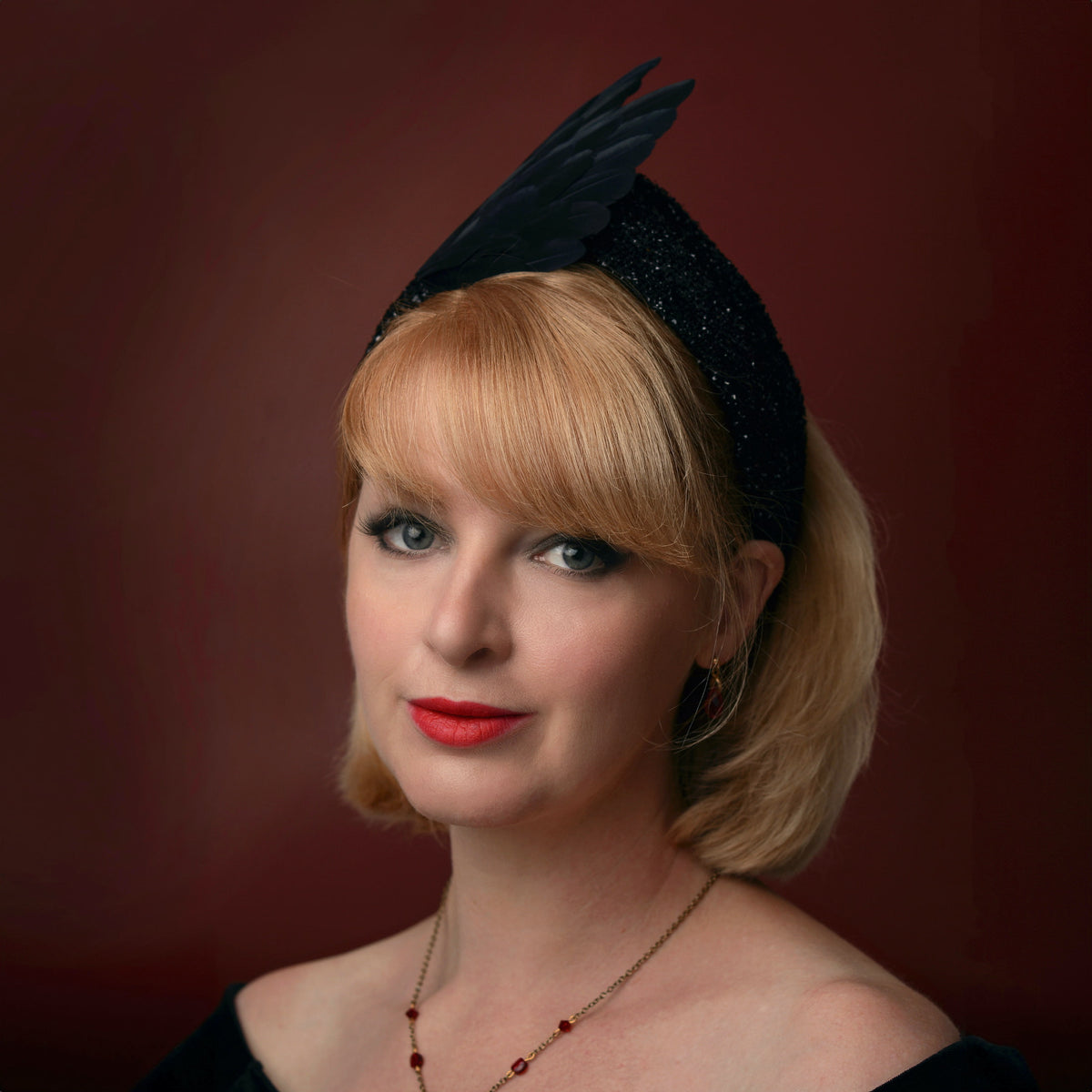 A woman with blonde hair wearing a black winged feathered fascinator headband, red lipstick, and a delicate necklace, set against a dark reddish-brown studio background.