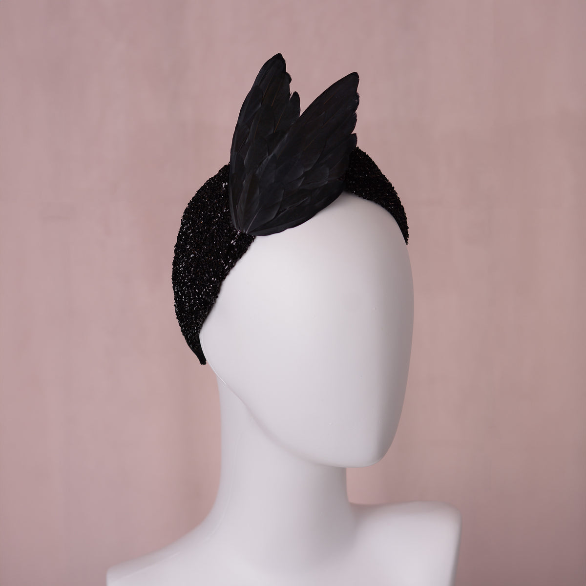 A black bird-wing feather fascinator headpiece with a glittery black fabric headband, displayed on a white mannequin head against a soft pink background.