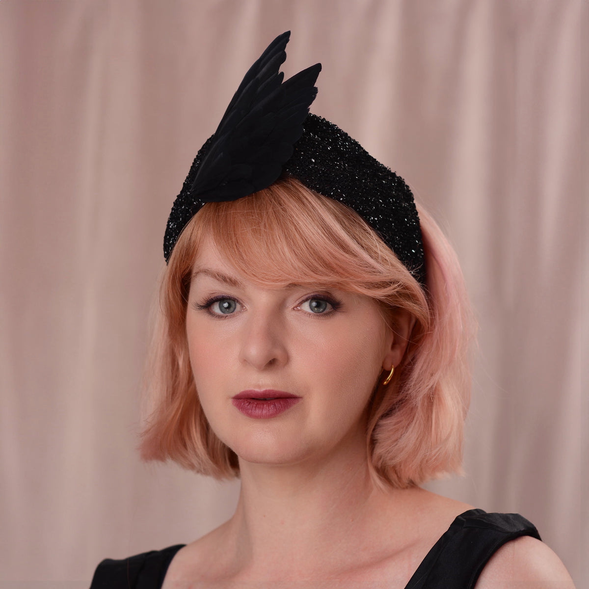 Woman with and soft pink bob haircut  wears a sparkling turban style headband in sparkling black fabric with black feather bird wings arranged in a v shape.