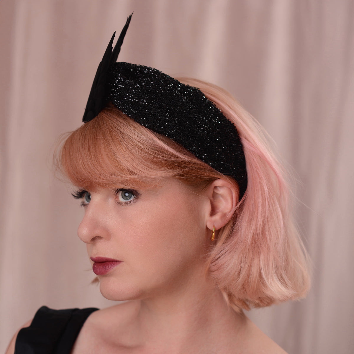 A woman with short pink hair models a black glitter fascinator headband with two upward-pointing feather  accents, set against a soft beige curtain background.