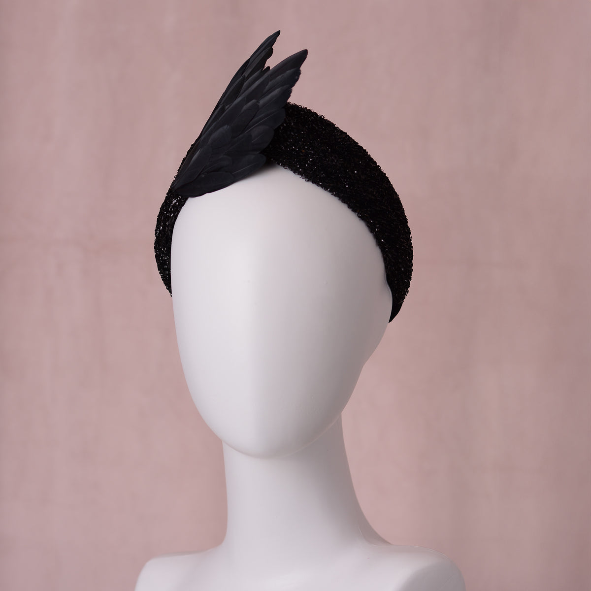 A black feather bird wing fascinator headpiece on a glitter black twist turban headband, displayed on a white mannequin head against a soft pink background.