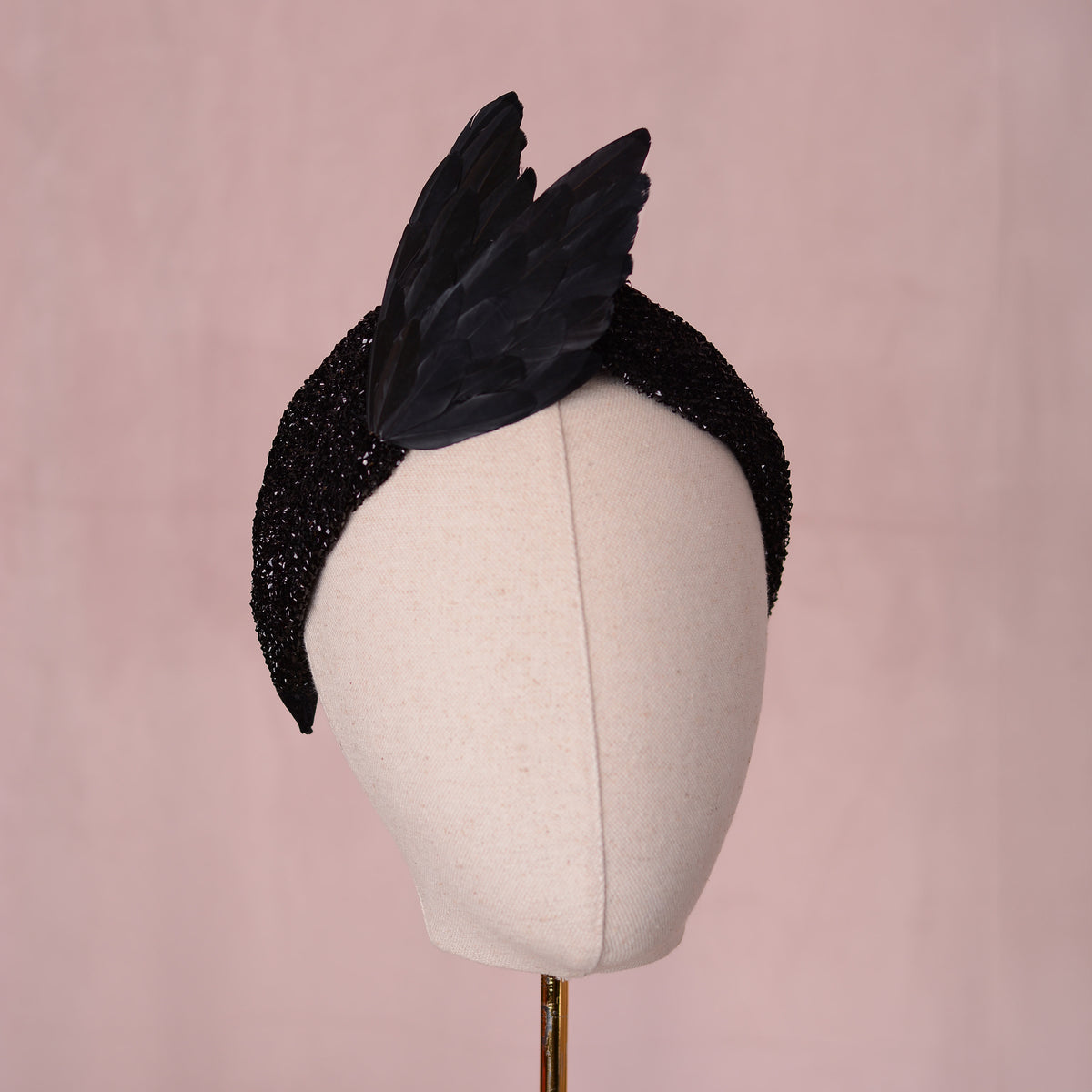 A black glitter fabric fascinator headpiece with two large black feathered bird wings in a elegant V shape. Displayed on a cream fabric mannequin head against a soft pink background.