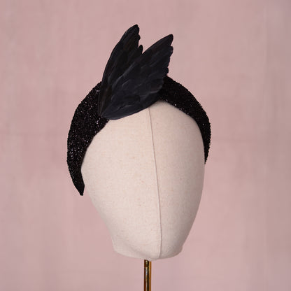 A black glitter fabric fascinator headpiece with two large black feathered bird wings in a elegant V shape. Displayed on a cream fabric mannequin head against a soft pink background.