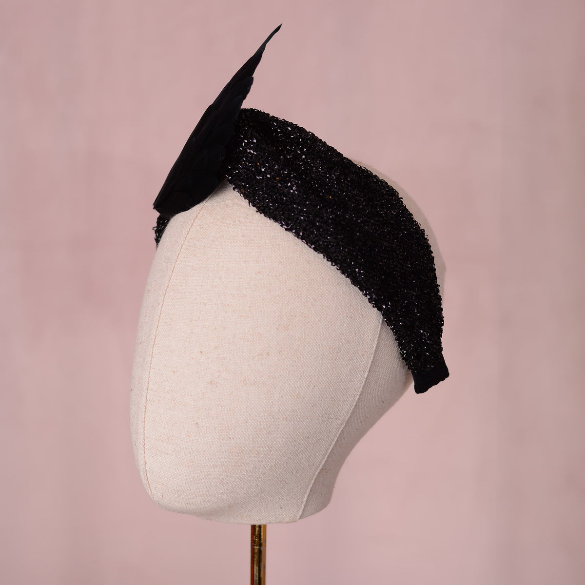 A gothic black embellished fascinator headpiece with two large black wings crafted in feathers, displayed on a cream-colored mannequin head against a soft pink background.