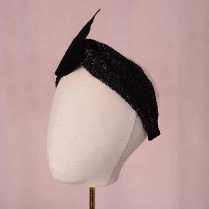 A gothic black embellished fascinator headpiece with two large black wings crafted in feathers, displayed on a cream-colored mannequin head against a soft pink background.