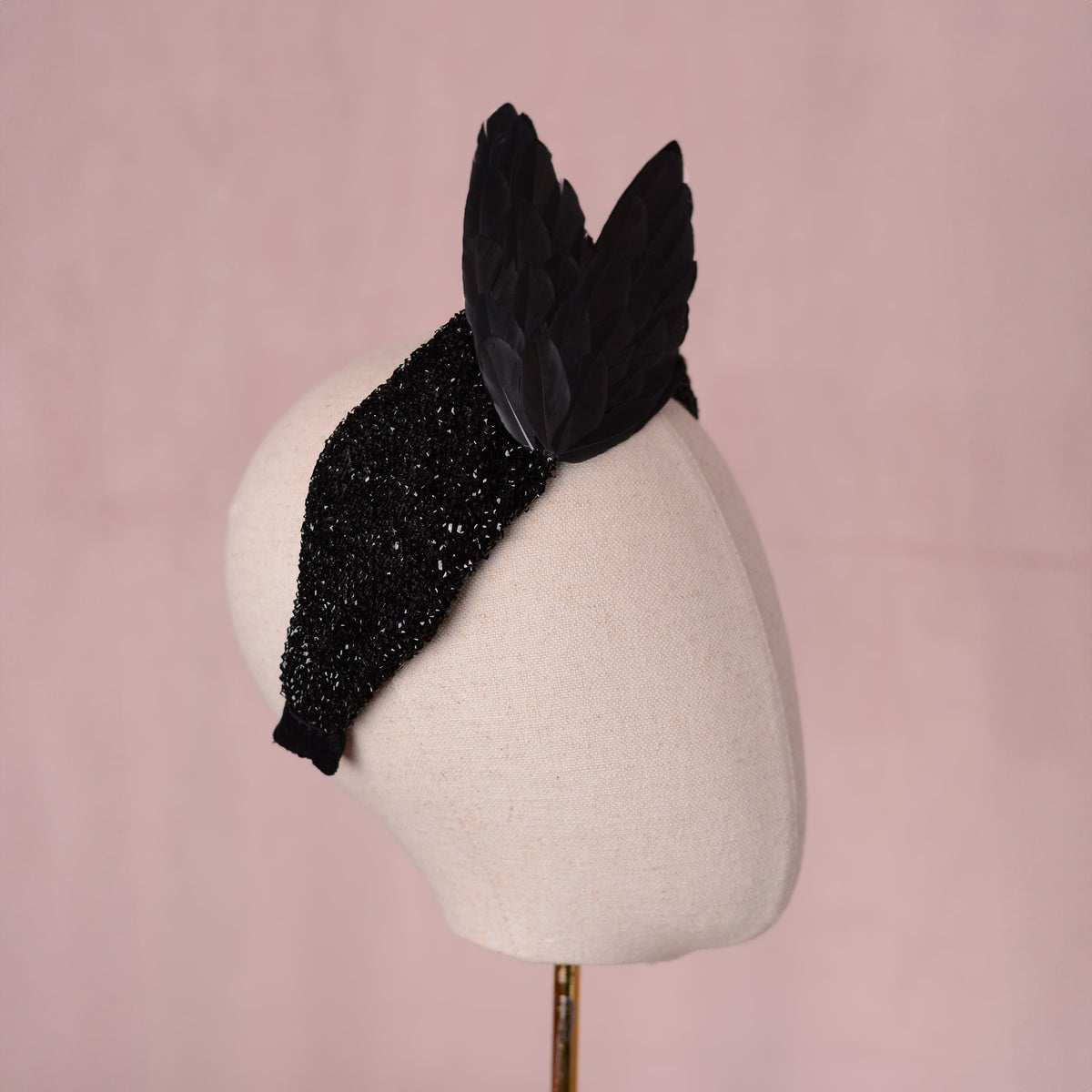 A gothic black embellished fascinator headpiece with two large black wings crafted in feathers, displayed on a cream-colored mannequin head against a soft pink background.