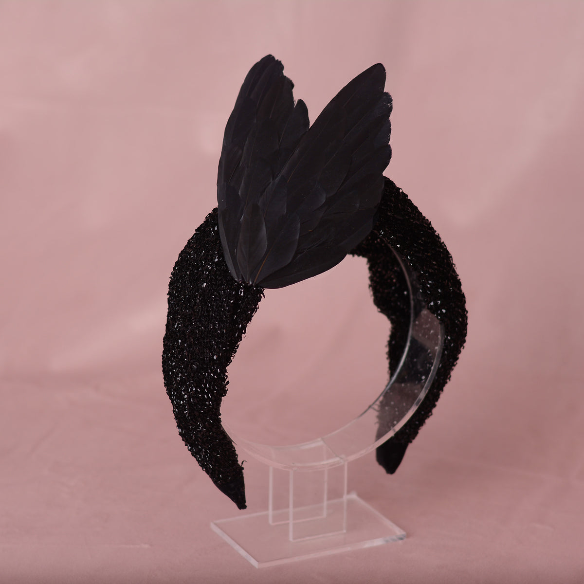 A black bird wing feather headband with a glittery black fabric headband base, displayed on a clear acrylic stand against a soft pink background.