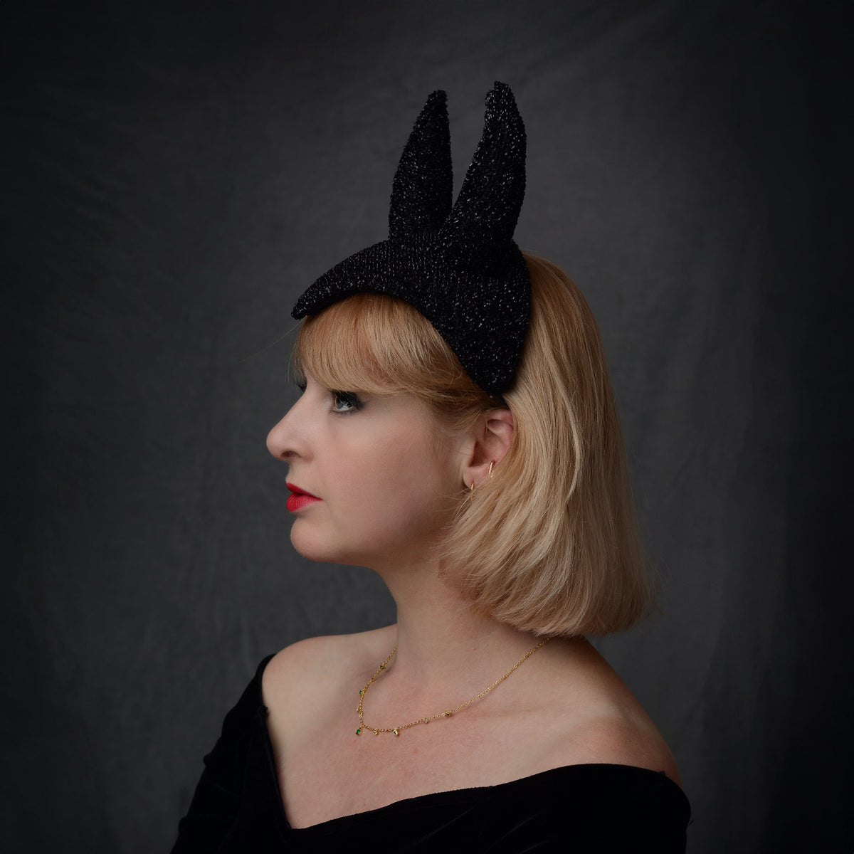 A woman in profile wearing a black glitter devil headpiece in a vintage show girl style against a dark gray studio background.