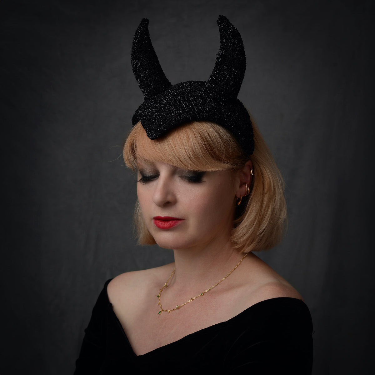 A woman with short blonde hair wearing a black glittery horned halloween themed headpiece, styled with a black off-the-shoulder top and a delicate gold necklace, set against a dark gray studio background.
