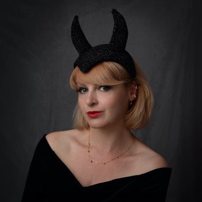 A woman with blonde hair wearing a black glitter devil horn headband, a black off-the-shoulder top, and a delicate gold necklace, set against a dark gray studio background.