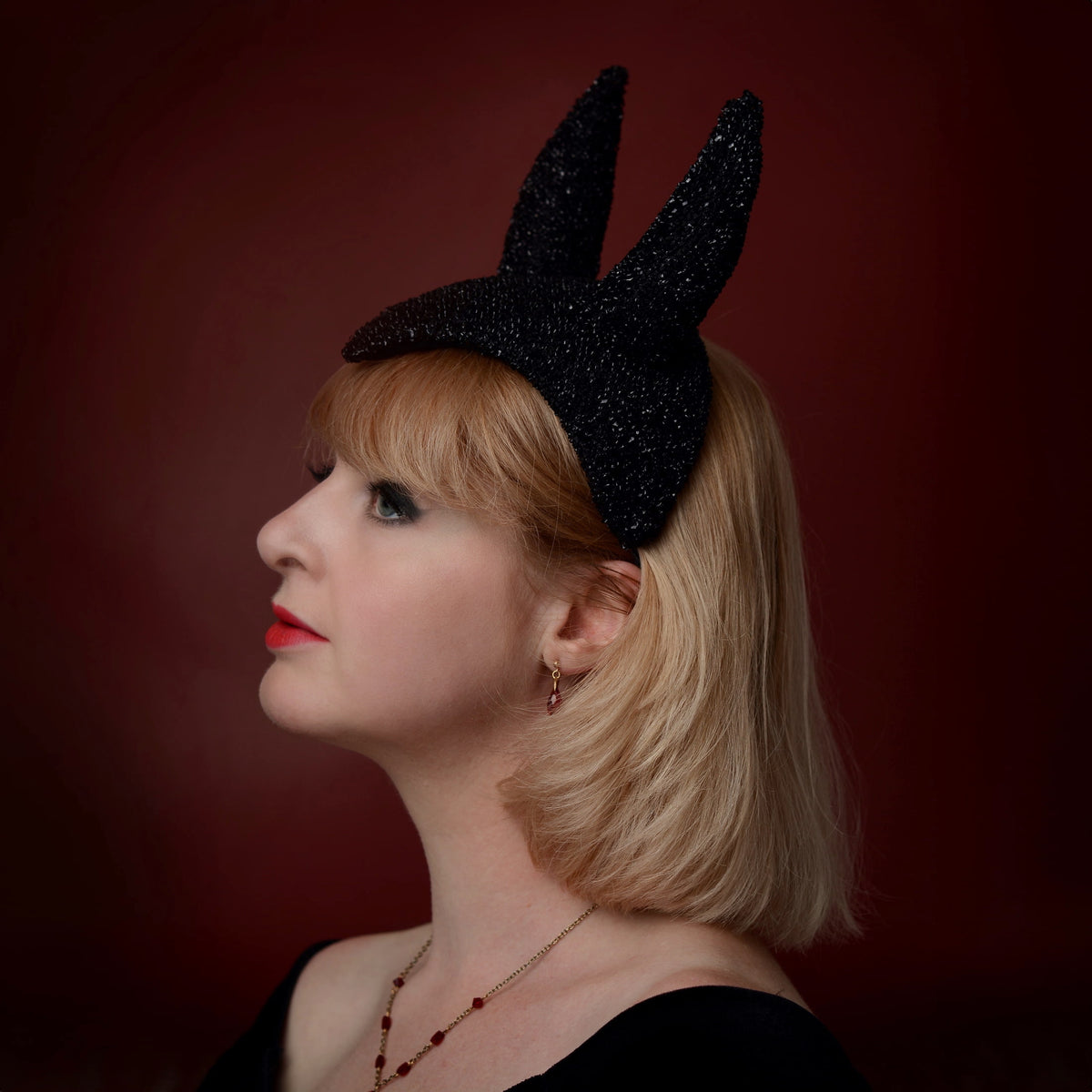 A woman in profile wearing a black glittery devil horn theatrical headpiece, set against a dark red studio background.