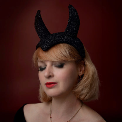 A woman with short blonde hair wearing a black glittery devil horn hat- a grown up glamourous halloween headpiece posed against a dark red background.