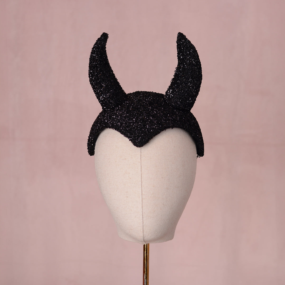 A black glittery devil horn headpiece displayed on a cream-colored mannequin head against a soft pink background.