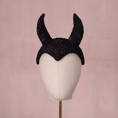 A black glittery devil horn headpiece displayed on a cream-colored mannequin head against a soft pink background.