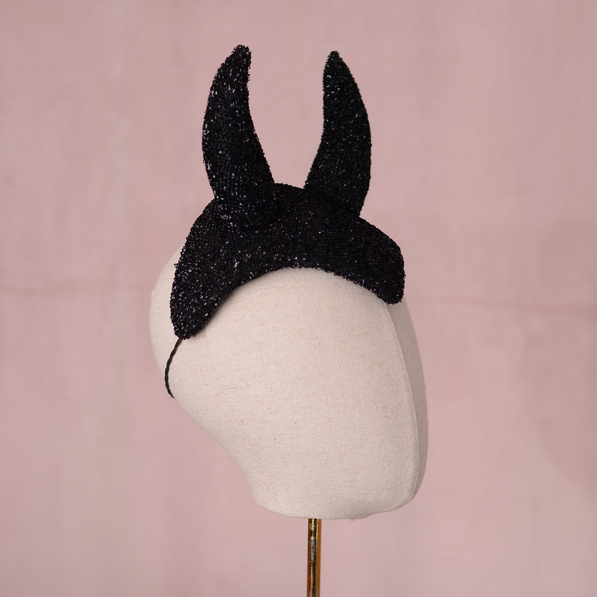 A black glitter fascinator headpiece with two pointed devil horns, displayed on a cream fabric mannequin head against a soft pink background.