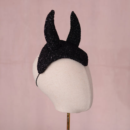 A black glitter fascinator headpiece with two pointed devil horns, displayed on a cream fabric mannequin head against a soft pink background.