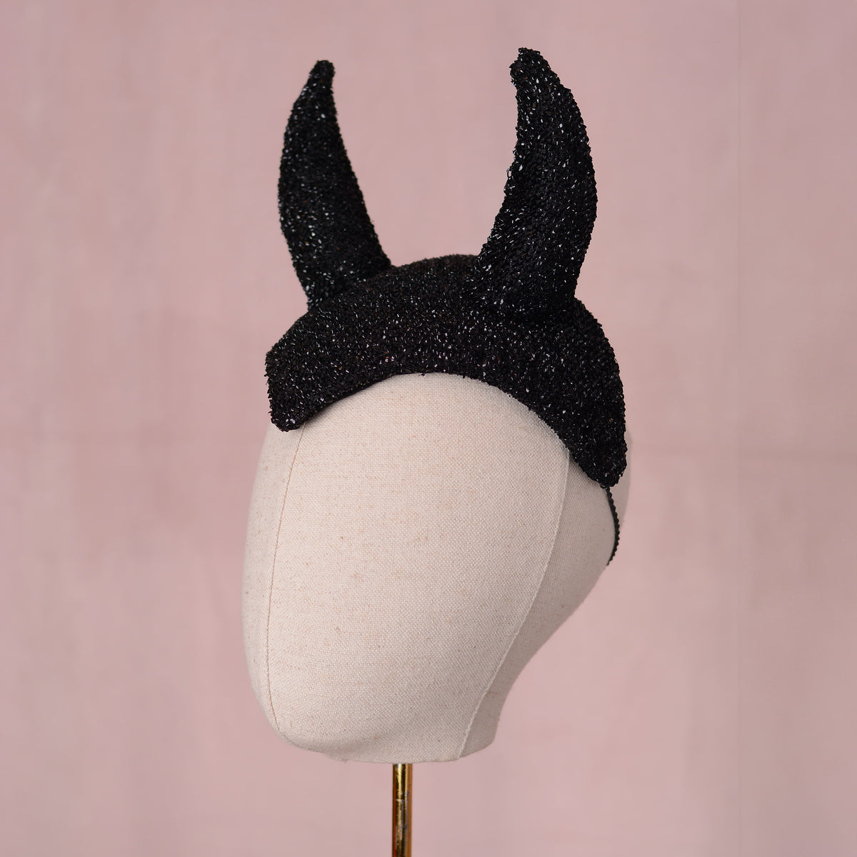 A black shimmering theatrical headpiece with two upright pointed horns, displayed on a cream fabric mannequin head against a soft pink background.
