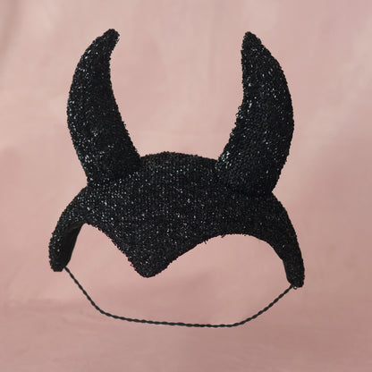 A black glittering devil horn headpiece  in a vintage show girl style with a thin black elastic hat band, displayed against a solid dusty pink background.