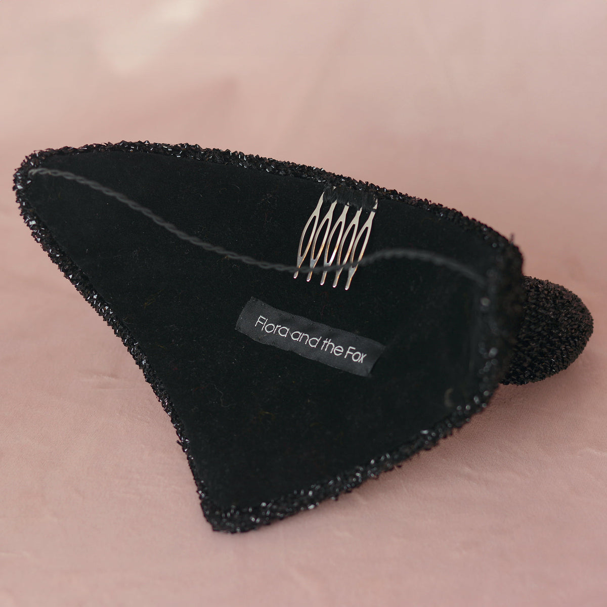 A black devil hat lined in velvet with a hat elastic and a metal comb featuring a fabric label that reads 'Flora and the Fox', set against a soft pink background.